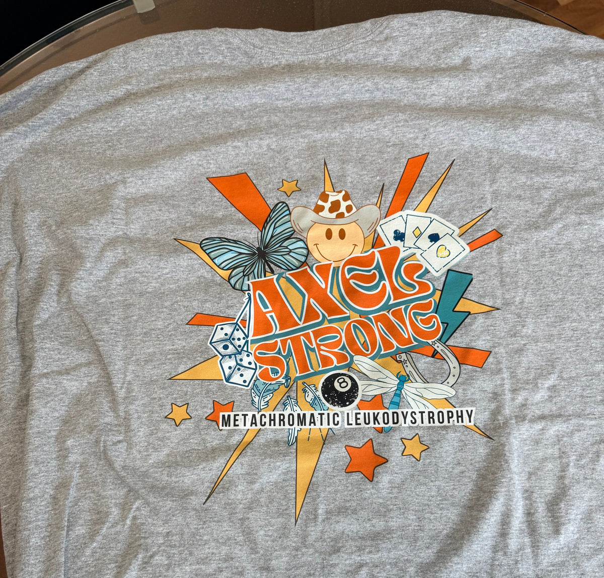 A heather gray short-sleeve t-shirt with a colorful graphic on the front featuring the text 'AXELSTRONG' and 'METACHROMATIC LEUKODYSTROPHY' along with various illustrative elements like a butterfly, stars, dice, and an 8-ball. 