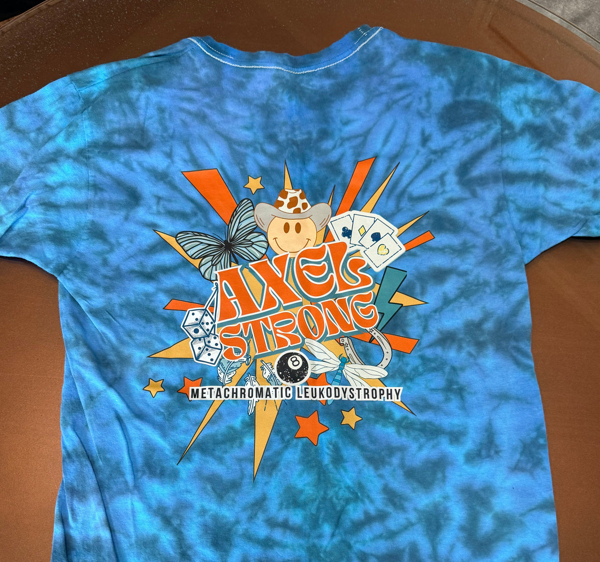 A blue tie-dye t-shirt with a large graphic on the back featuring the text 'AXELSTRONG' and 'METACHROMATIC LEUKODYSTROPHY' with various colorful retro illustrations. 