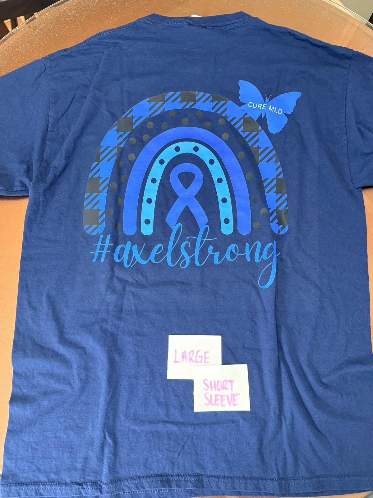 A blue short-sleeve t-shirt laid flat on a tan surface, showing the back side with a large graphic design. The design features a blue awareness ribbon inside a rainbow made of blue and dark plaid sections, a blue butterfly, and the text 'CURE MD' and '#axelstrong'.