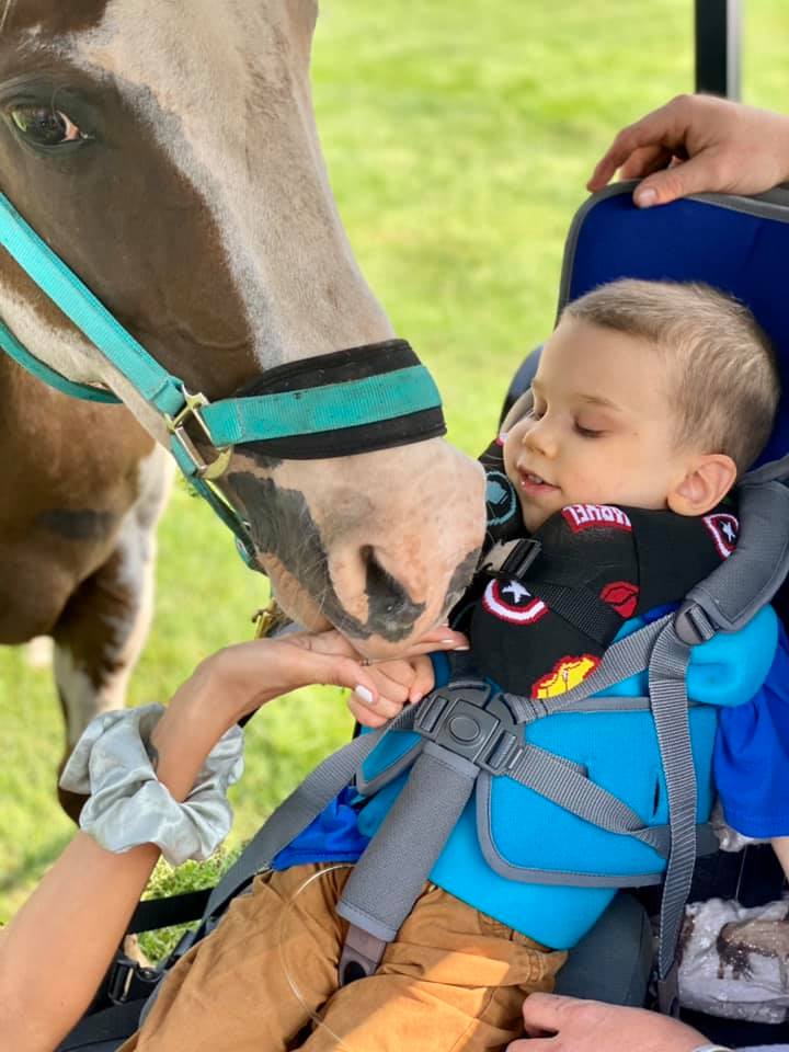 A young child wearing a bright blue and gray horse carrier seat sits on a blue saddle while a horse gently licks the child's hand. The child is also wearing a black Captain America themed neck pillow.