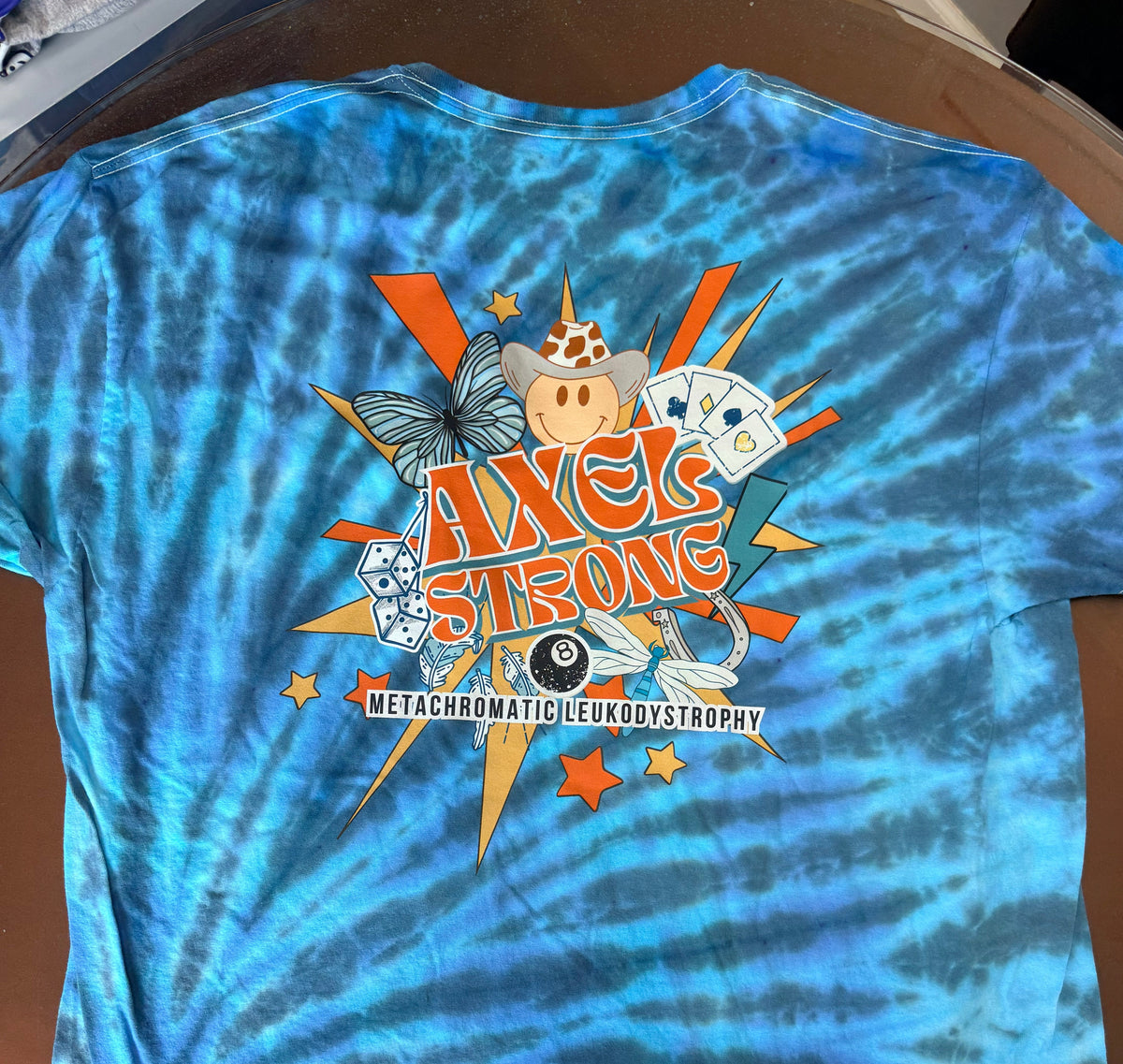 A blue and teal tie-dye t-shirt with a large graphic on the back featuring the text 'AXELSTRONG' and 'METACHROMATIC LEUKODYSTROPHY' with various mental health icons. 