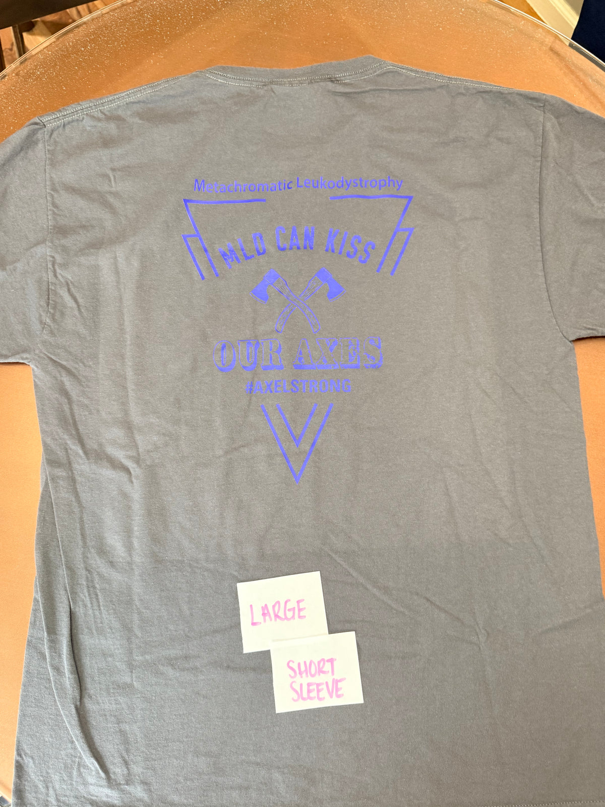 A gray short-sleeve t-shirt laid flat on a wooden surface, showing the back side with a blue graphic print that reads 'MACE CAN RIDE' and 'OUR AXES FUTURE STRONG'. Two small white sticky notes are placed on the lower left of the shirt.