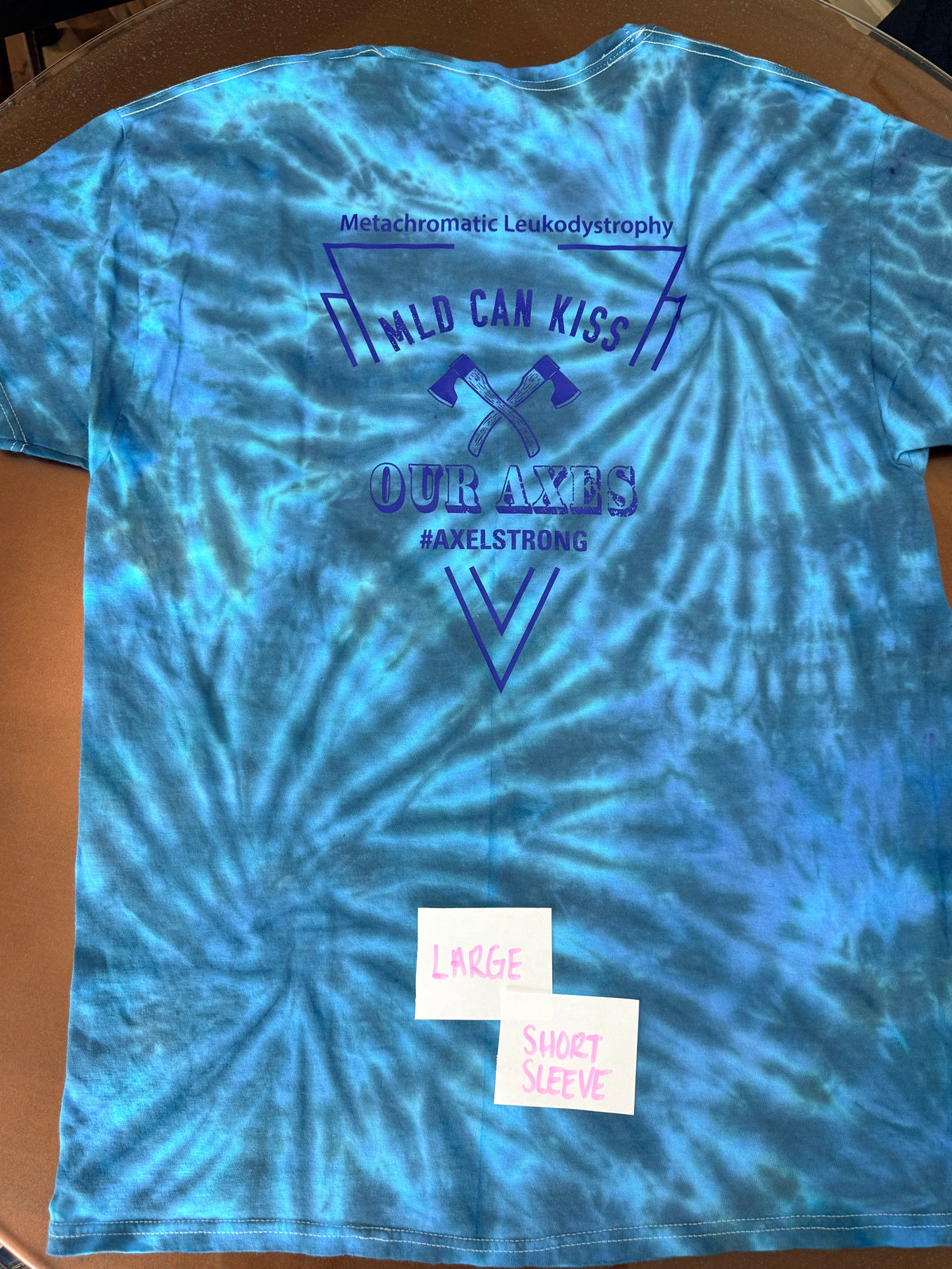 A blue and teal tie-dye t-shirt with a dark blue graphic print on the front. The graphic features the text 'Metachromatic Leukodystrophy', 'MILD CAN KISS', 'OUR AXES', and '#AXELSTRONG' with crossed axes.