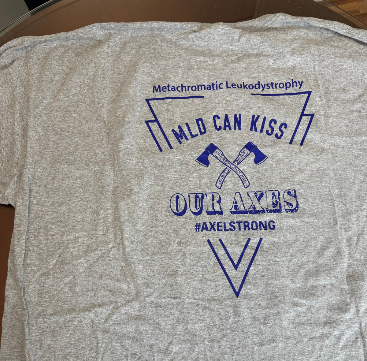 A heather gray short-sleeve t-shirt laid flat, featuring a blue graphic print on the front that reads 'Metachromatic Leukodystrophy', 'MILD CAN KISS', 'OUR AXES', and '#AXELSTRONG' with crossed axes. Two small handwritten size tags reading 'SHORT SLEEVE' and '3YL' are placed near the bottom.