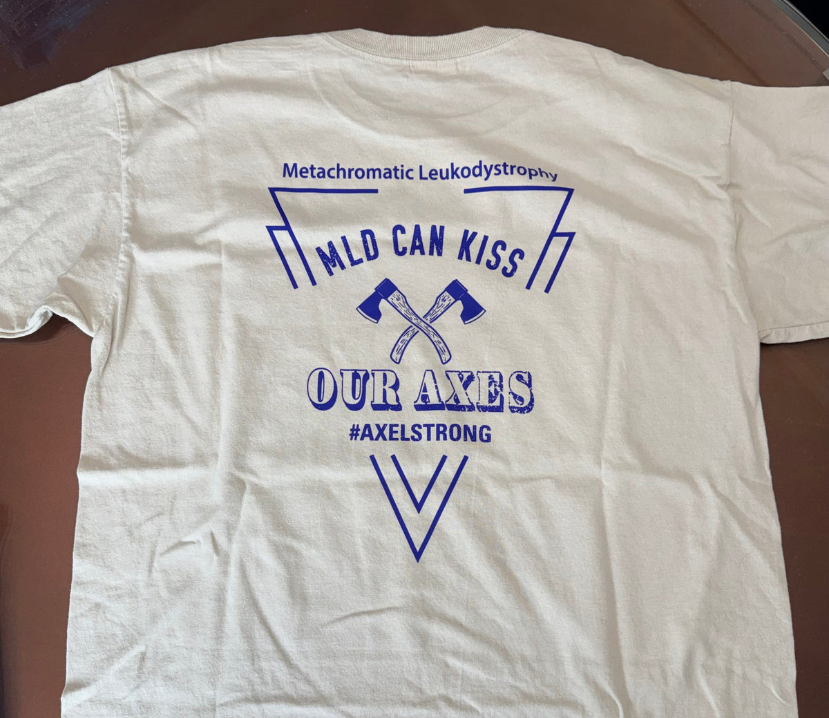 A cream-colored short-sleeve t-shirt laid flat on a dark brown surface, showing the back graphic with blue text and illustrations. 