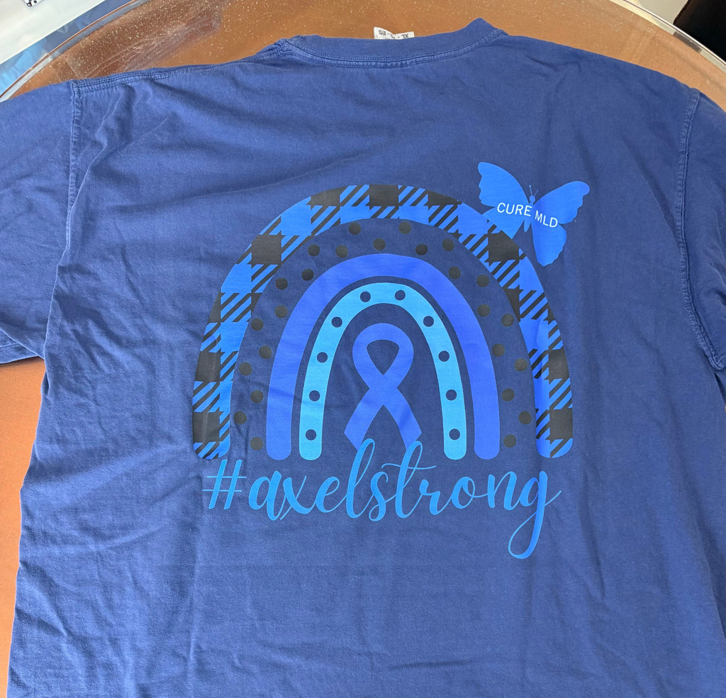 A blue short-sleeve t-shirt laid flat on a wooden surface, showing the back with a large graphic of a rainbow, awareness ribbon, butterfly, and the text '#axelstrong'. 