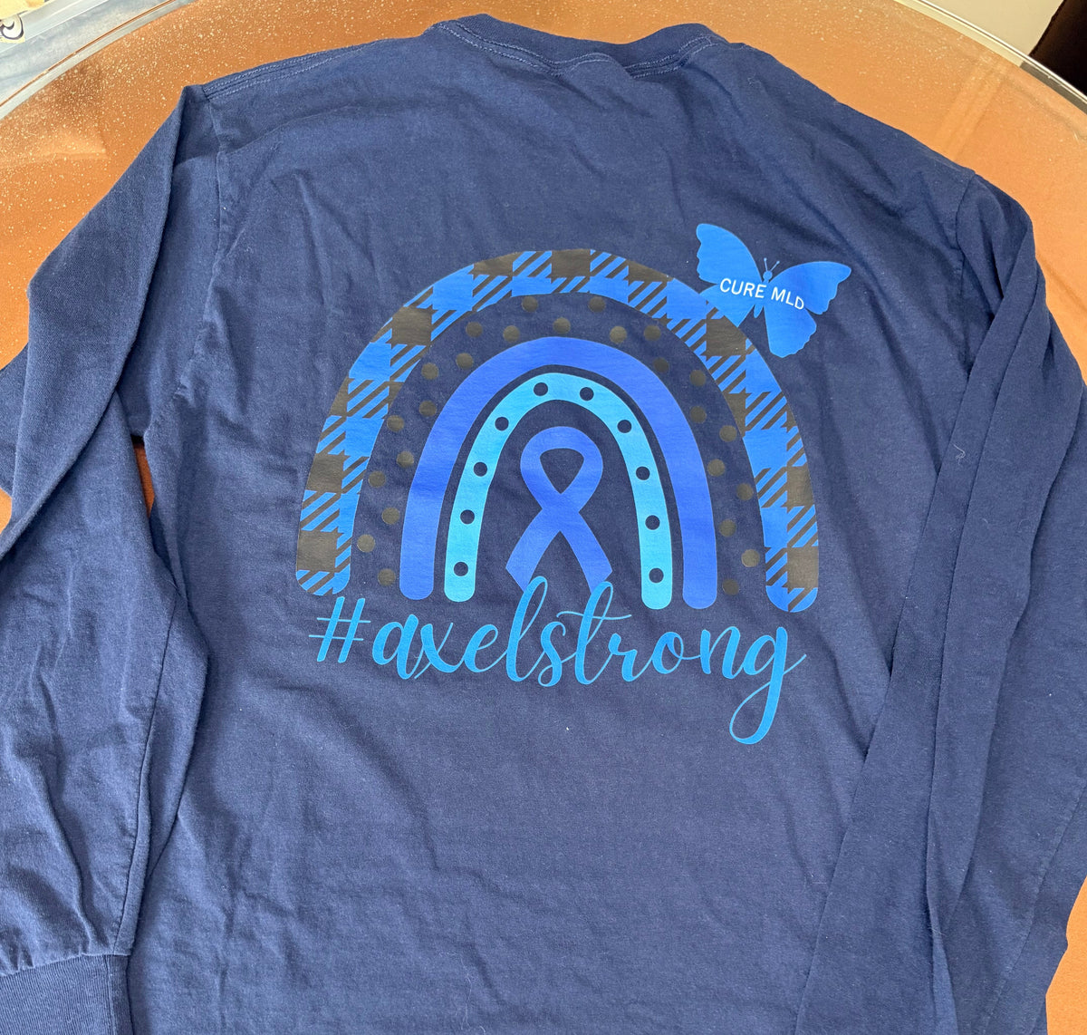 A navy blue long-sleeve shirt laid flat on an orange surface, showing the back with a large blue and gray awareness-themed rainbow graphic, a ribbon motif, a butterfly, and the text '#axelstrong'. 
