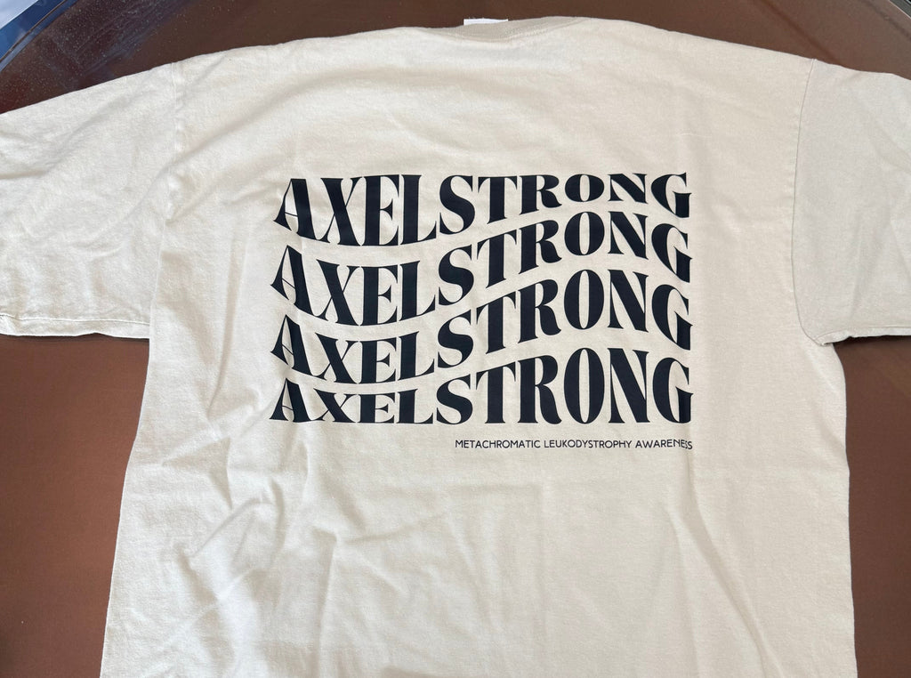 A cream-colored short-sleeve t-shirt laid flat on a brown glass table, featuring the word 'AXELSTRONG' repeated in a wavy black font across the chest. 