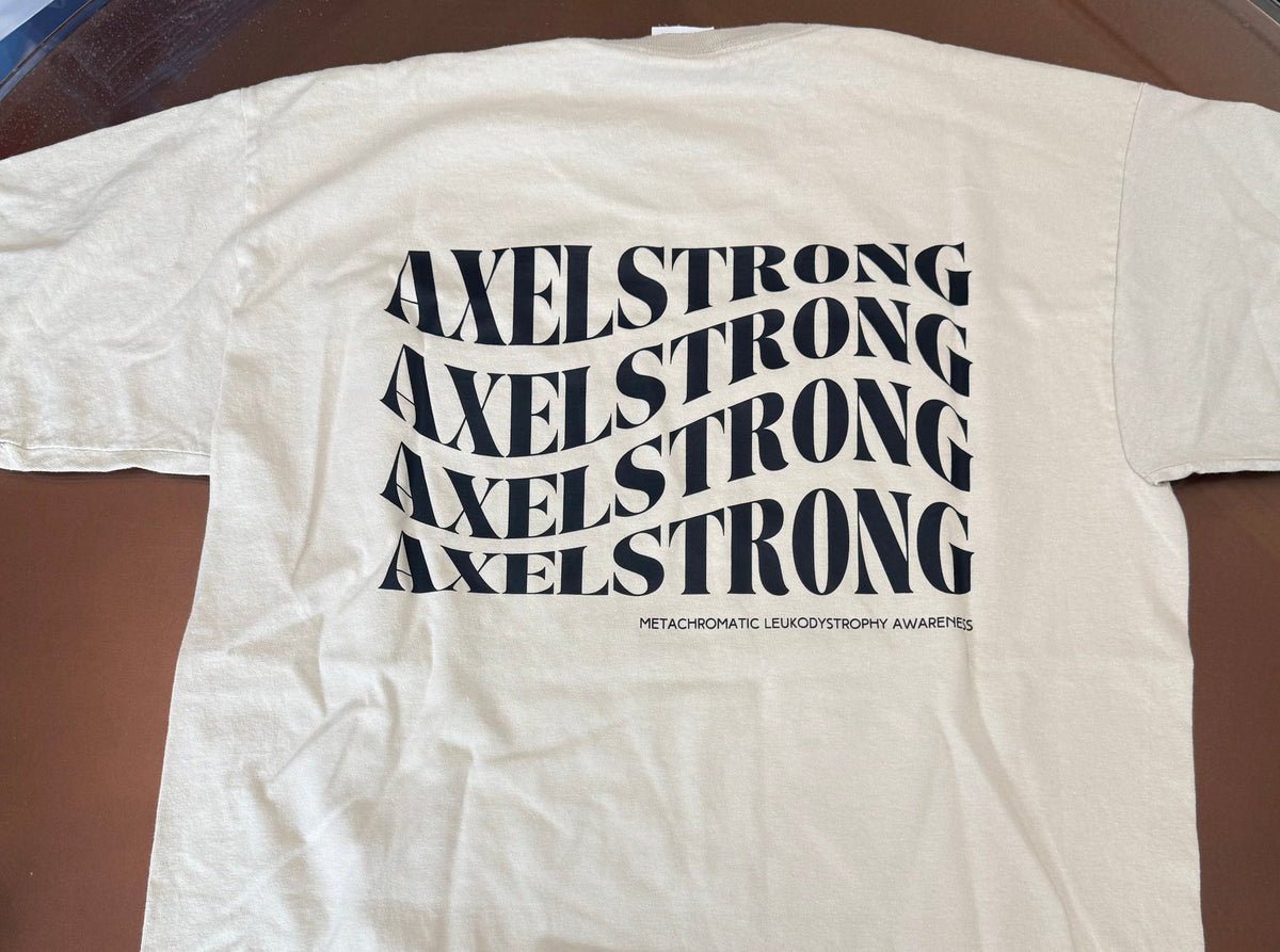 A cream-colored short-sleeve t-shirt laid flat on a brown glass table, featuring the word 'AXELSTRONG' repeated in a wavy black font across the chest. 