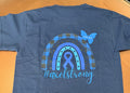 A blue short-sleeve T-shirt laid flat on an orange surface, showing the back with a large rainbow and awareness ribbon graphic in blue and gray, plus the text '#axelstrong'. 