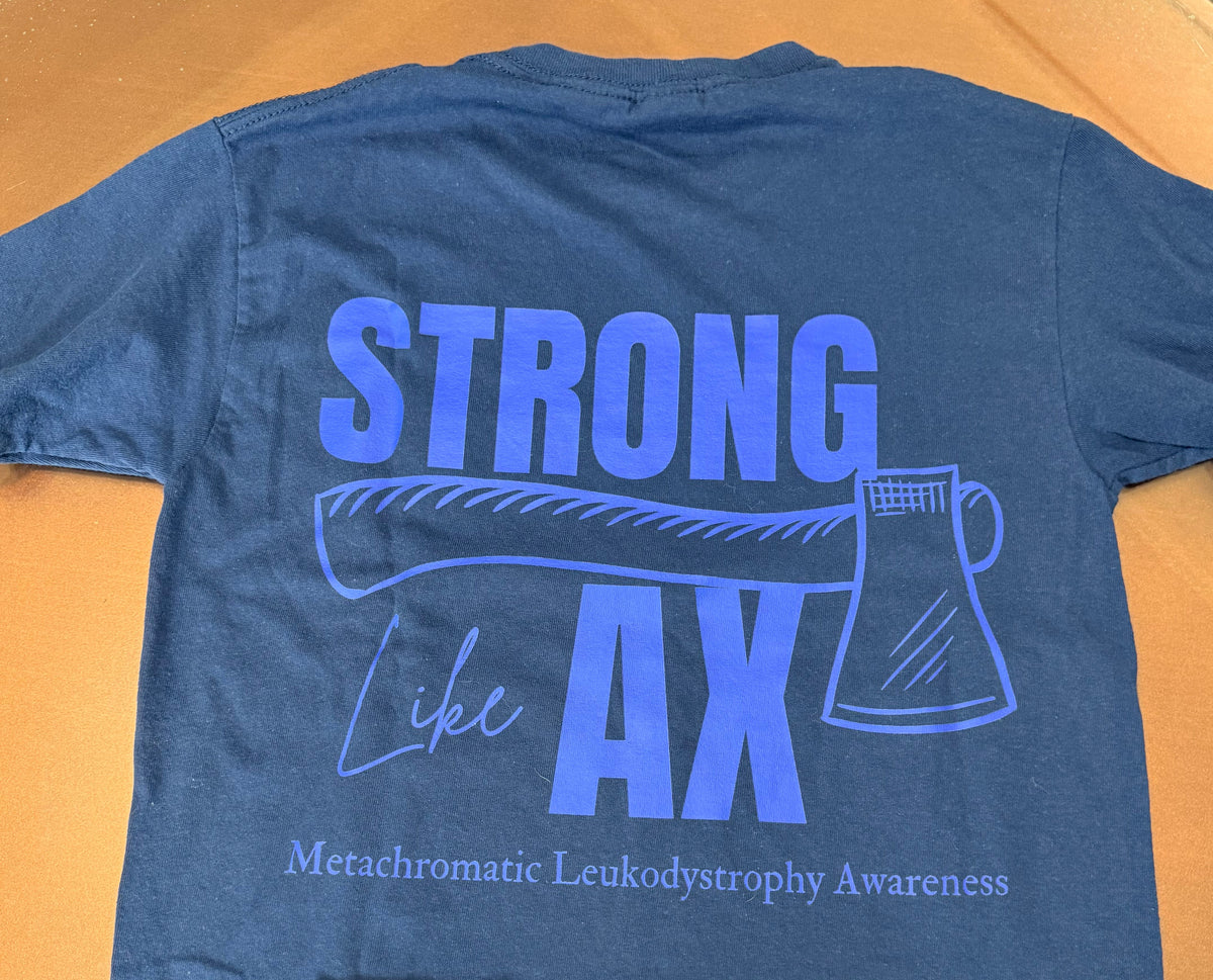 A blue short-sleeve t-shirt laid flat on an orange-brown surface, showing the back with a large light blue graphic that reads “STRONG LIKE AX” and “Metachromatic Leukodystrophy Awareness.” 