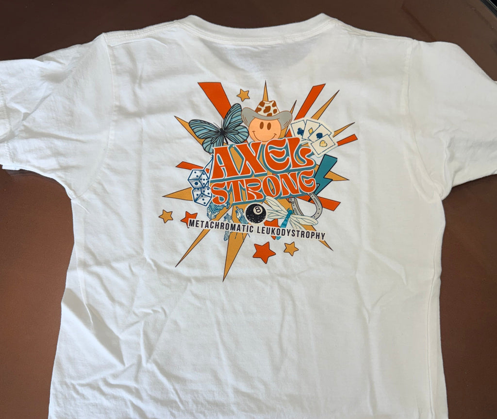 A white short-sleeve t-shirt laid flat on a brown surface, featuring a colorful retro-style graphic on the chest with the text 'AXELSTRONG' and 'METACHROMATIC LEUKODYSTROPHY' surrounded by stars, a butterfly, and cartoon elements. 