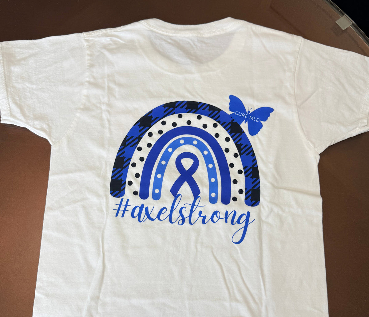 A white short-sleeve T-shirt laid flat on a dark brown surface, featuring a blue awareness ribbon and rainbow graphic on the front with the text '#axelstrong' and a blue butterfly. 